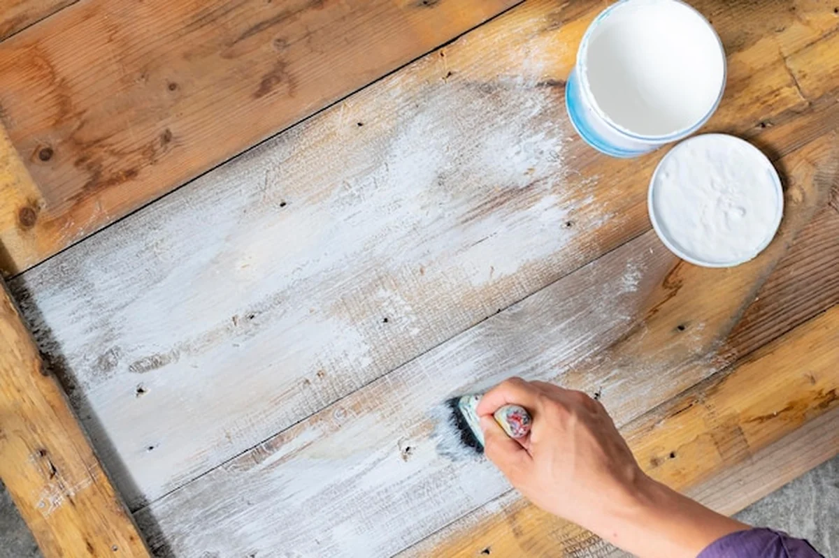 Dłoń malarza trzymająca pędzel, malowany drewniany stół na biało, biały słoik obok